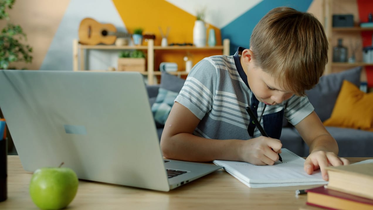 Enfant faisant ses devoirs devant un ordinateur portable et un cahier
